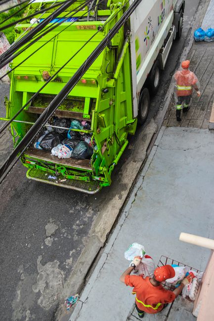 An overhead view of a bright green waste collection truck parked alongside a paved street, with its rear open and filled with black plastic rubbish bags, some transparent, containing assorted waste. T