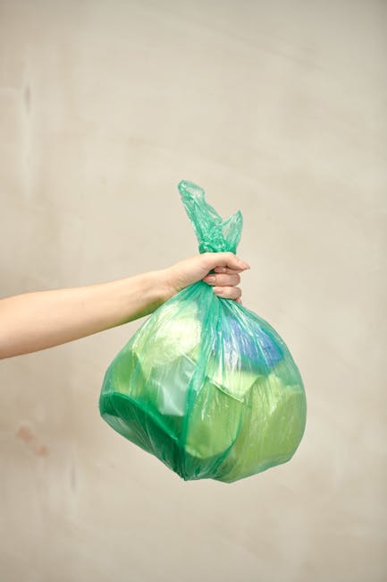 Two large black plastic rubbish bags, slightly crinkled and tied at the top, are placed on a paved sidewalk near a black metal fence. One bag appears to contain household waste or discarded packaging,